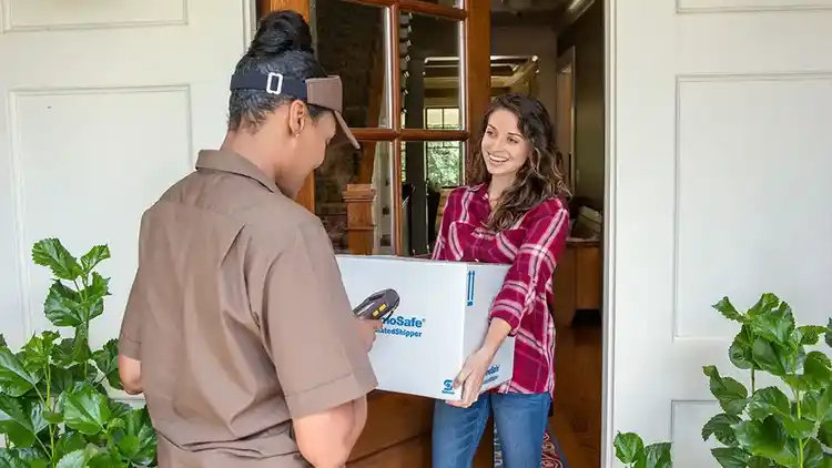 Eine Person an einer Haustür hält ein schweres Paket zum Versenden, das von einer UPS-Mitarbeiterin abgeholt wird