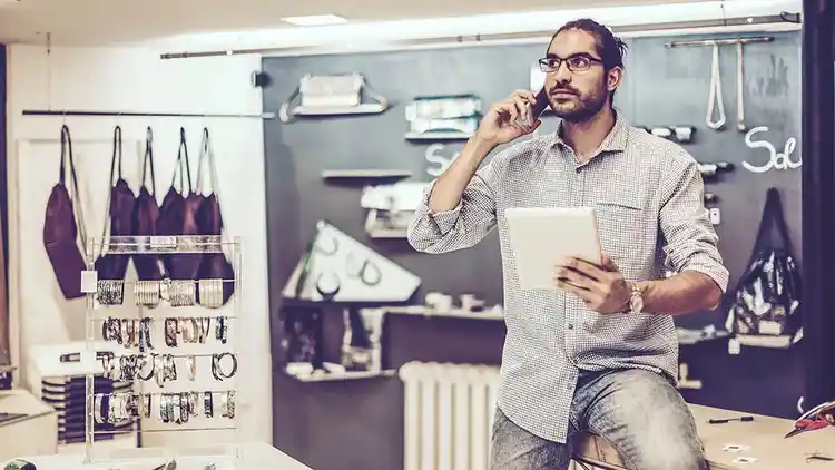 En man lutar sig mot ett skrivbord samtidigt som han pratar i en mobiltelefon och håller i en surfplatta
