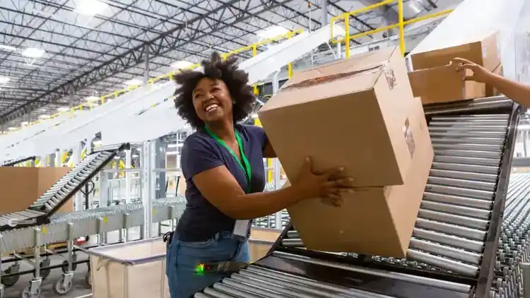 A grinning woman collecting boxes at the bottom of a sorting chute and pushing them along an extendable conveyor belt towards a truck loading dock for delivery
